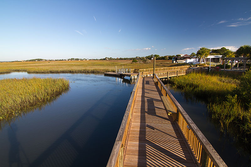 Wooden boardwalk extending over a calm marshland with grassy banks under a clear blue sky. Houses and small buildings are visible in the distant background, and sunlight casts strong shadows on the boardwalk.