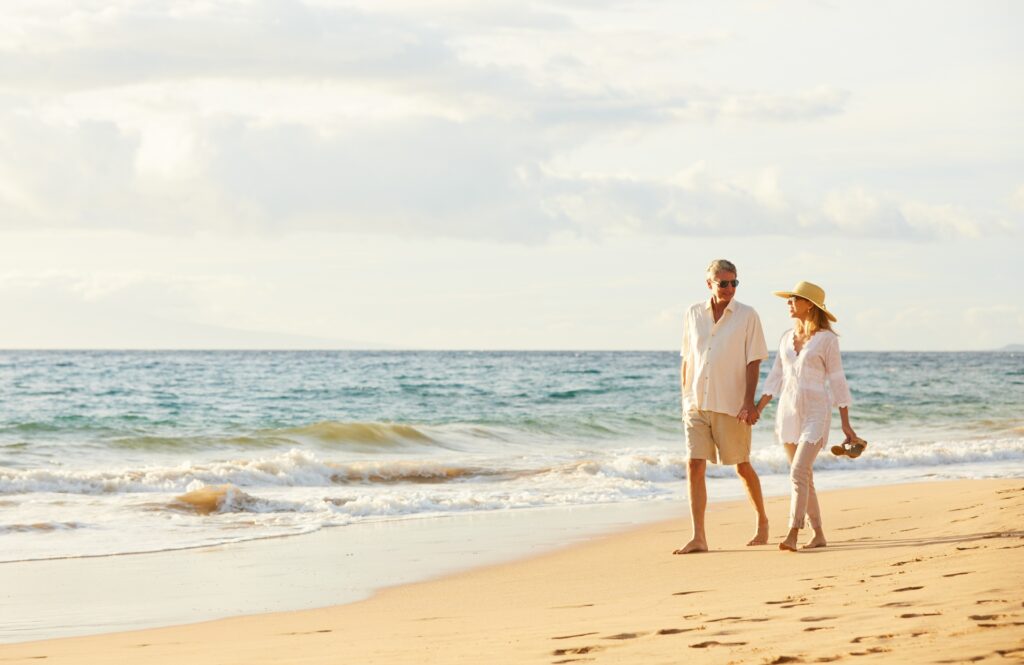 A senior couple of seasonal travelers walks hand in hand along a beach at sunset. The man is wearing a white shirt and beige shorts, while the woman is dressed in a white blouse and light pants with a sunhat. The serene ocean and soft waves are visible in the background.