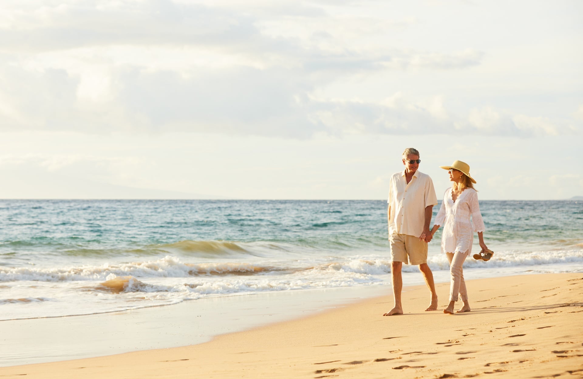 A senior couple of seasonal travelers walks hand in hand along a beach at sunset. The man is wearing a white shirt and beige shorts, while the woman is dressed in a white blouse and light pants with a sunhat. The serene ocean and soft waves are visible in the background.