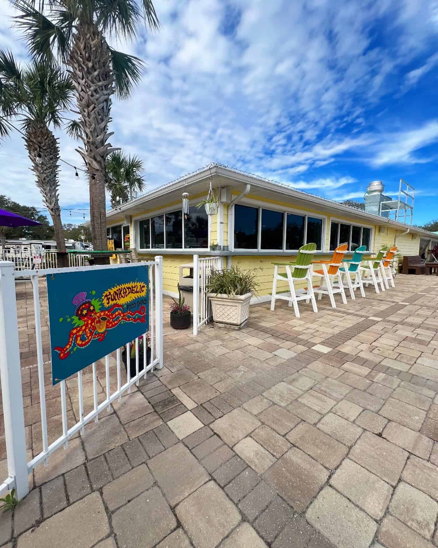 Exterior of a tropical-themed restaurant with palm trees, colorful high-back chairs, and a vibrant sign on the fence. The sky is partly cloudy, and the ground is paved with stone tiles.