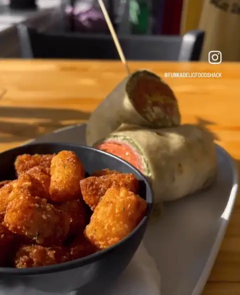 A bowl of crispy tater tots sits next to a wrap cut in half, filled with greens and red ingredients. A toothpick holds it together on a light-colored plate, placed on a wooden table. Sunlight casts soft shadows across the scene.