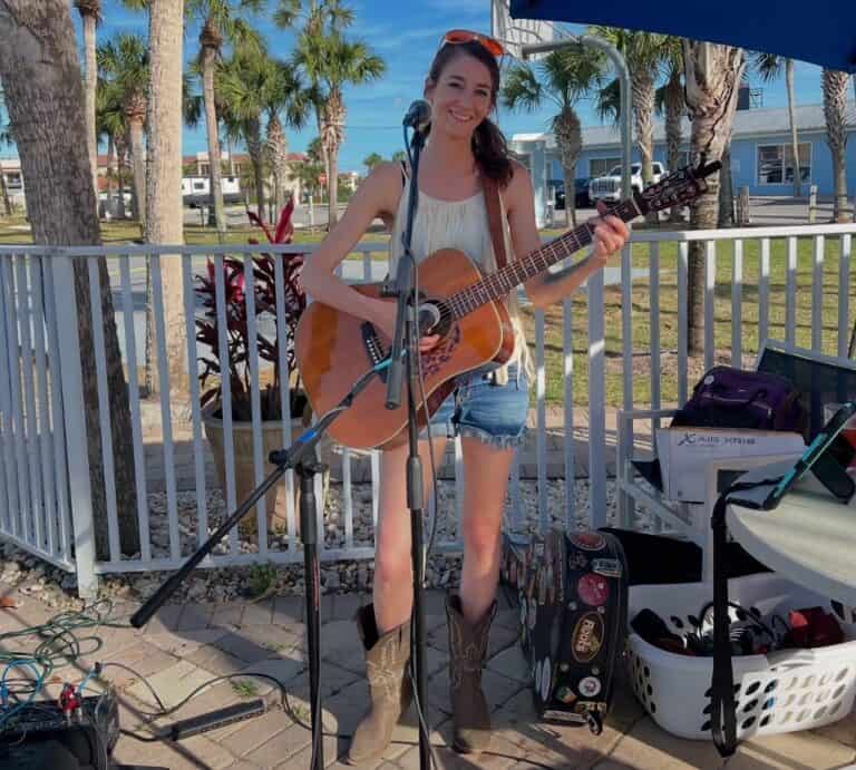 A woman stands smiling with a guitar, dressed in a sleeveless top, denim shorts, and boots. She is outdoors near palm trees and a fence, performing with microphones and equipment nearby. A sunny sky is visible in the background.
