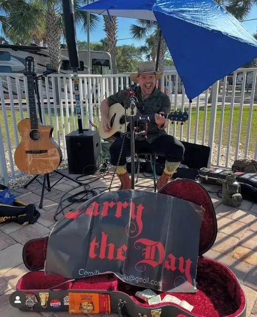 A musician sits under a blue umbrella, playing an acoustic guitar and singing into a microphone. Equipment and a second guitar surround him. A large sign reading "Carry the Day" is propped up in front. Palm trees are visible behind.