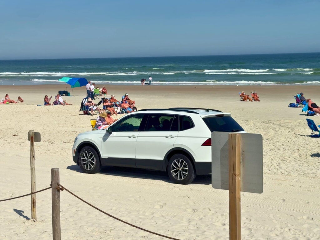 A white SUV is parked on a sandy beach with people sitting on chairs and towels near the ocean under a clear blue sky.
