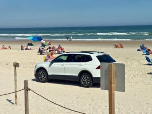 A white SUV is parked on a sandy beach with people sitting on chairs and towels near the ocean under a clear blue sky.