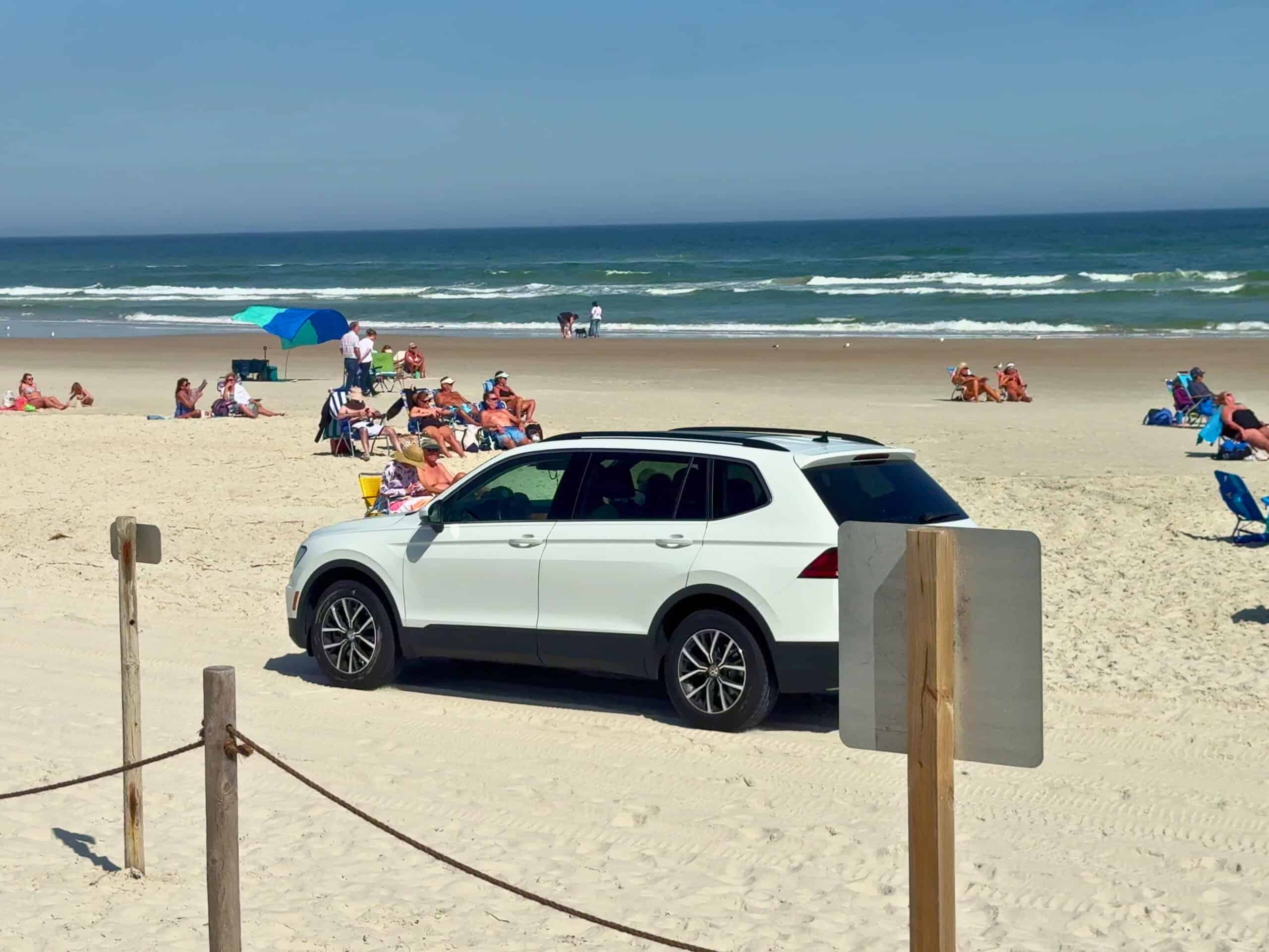 A white SUV is parked on a sandy beach with people sitting on chairs and towels near the ocean under a clear blue sky.