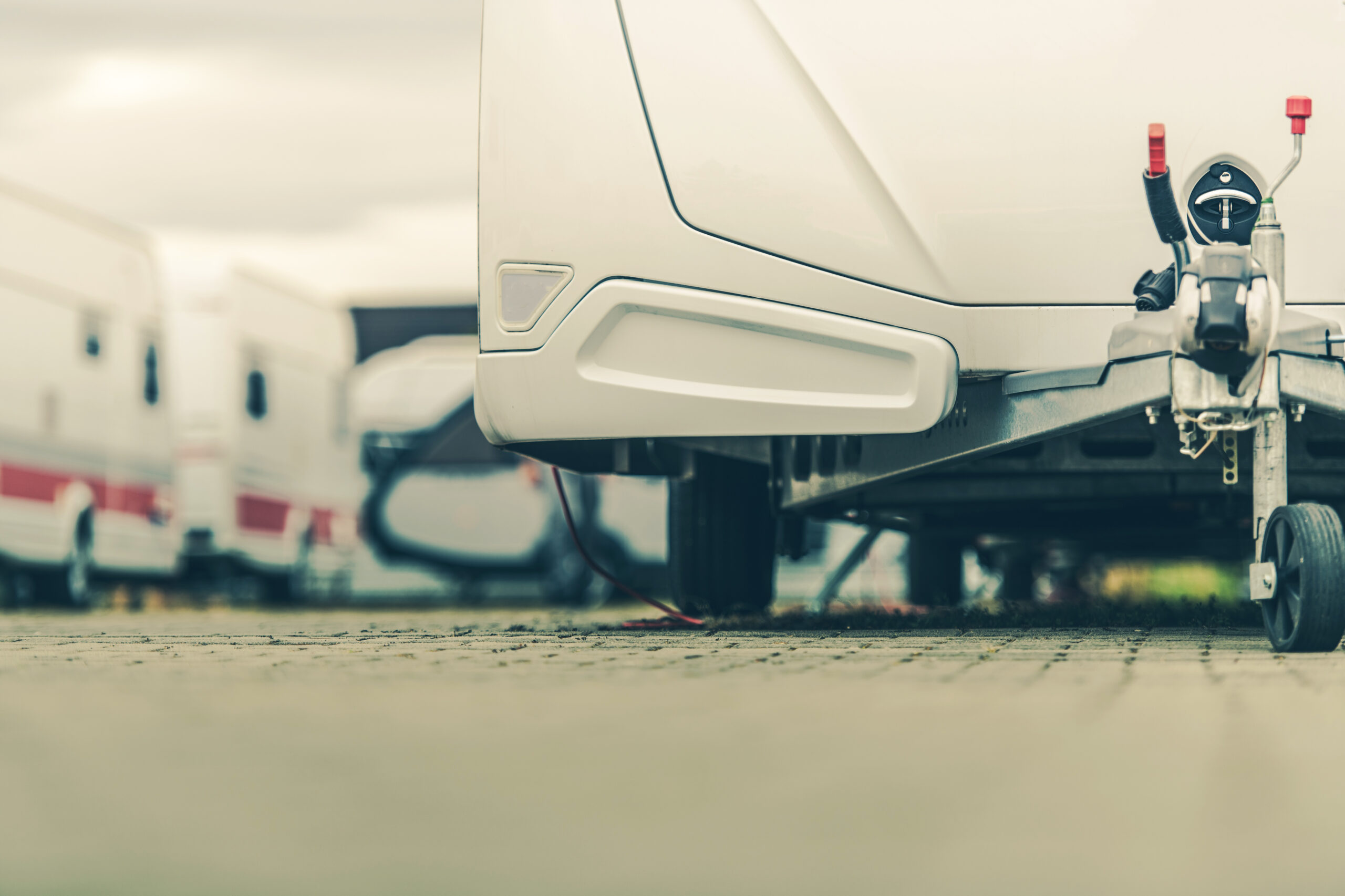 Low-angle view of a white travel trailer parked on a paved surface, with other trailers visible in the blurred background. The image focuses on the hitch and wheel area under the trailer.