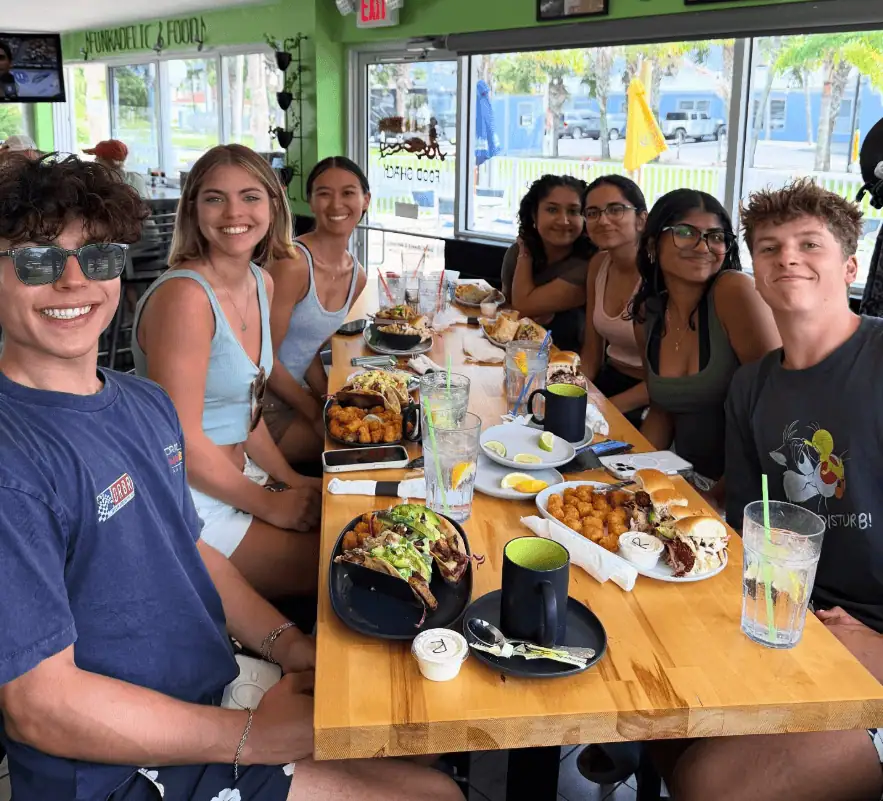 A group of seven young adults sit around a wooden table at a restaurant, smiling at the camera with plates of food and drinks in front of them.