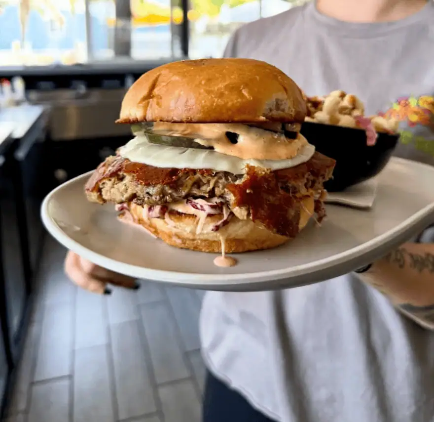 A person holds a plate with a sandwich topped with sliced onions, pickles, sauce, and coleslaw, with a side dish in the background.