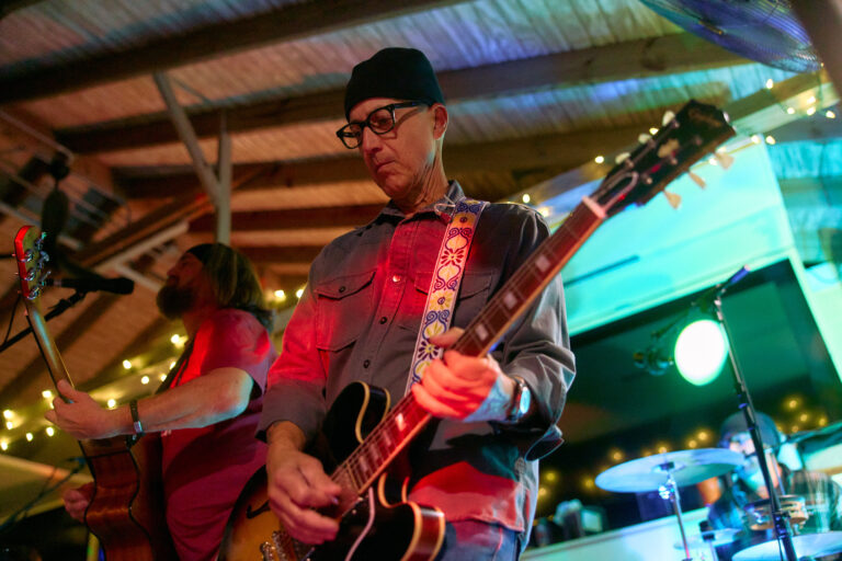 Two musicians playing electric guitars on stage under string lights, with a drummer visible in the background.