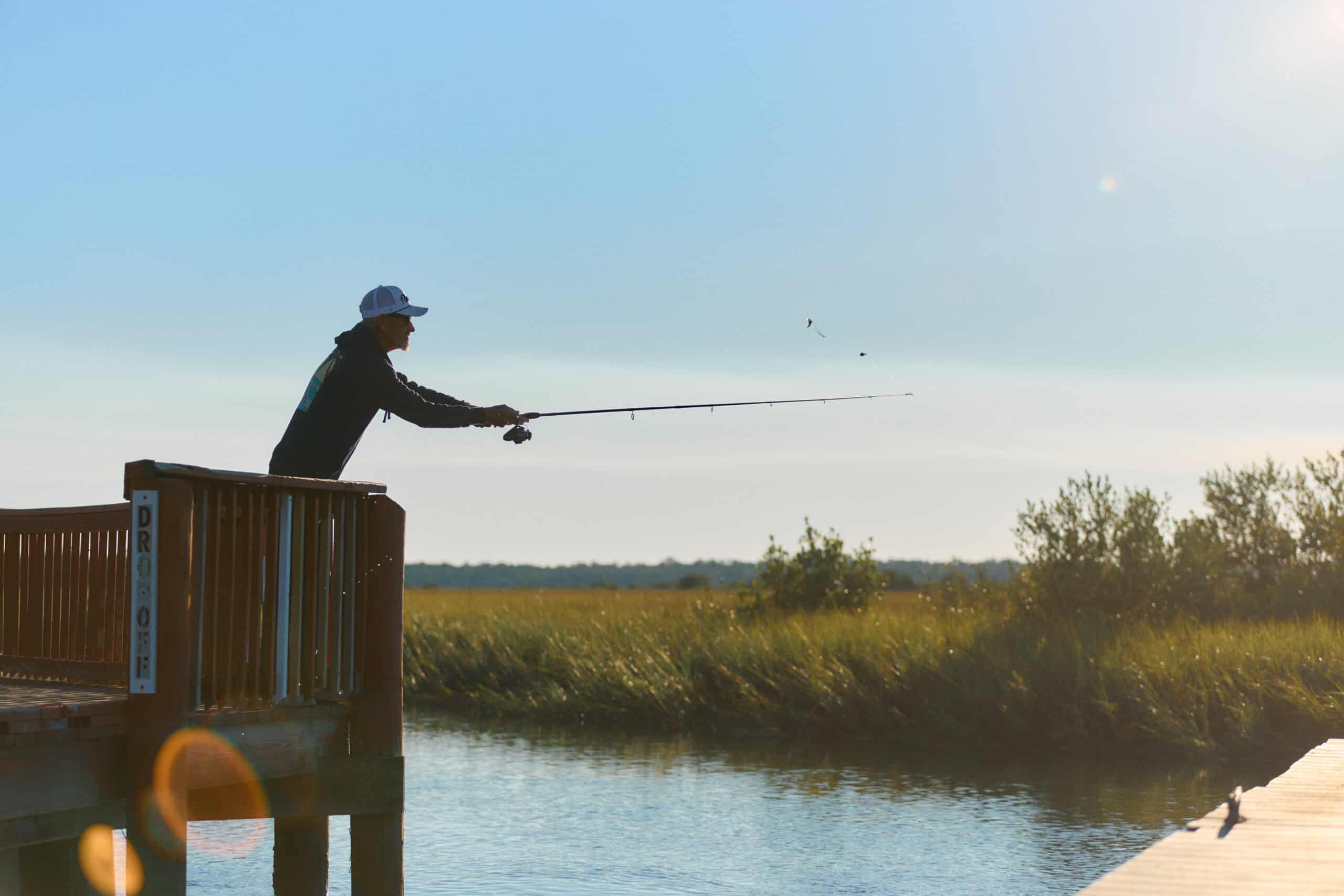 A person in a cap casts a fishing line from a wooden dock into a calm body of water, with tall grass and clear sky in the background.