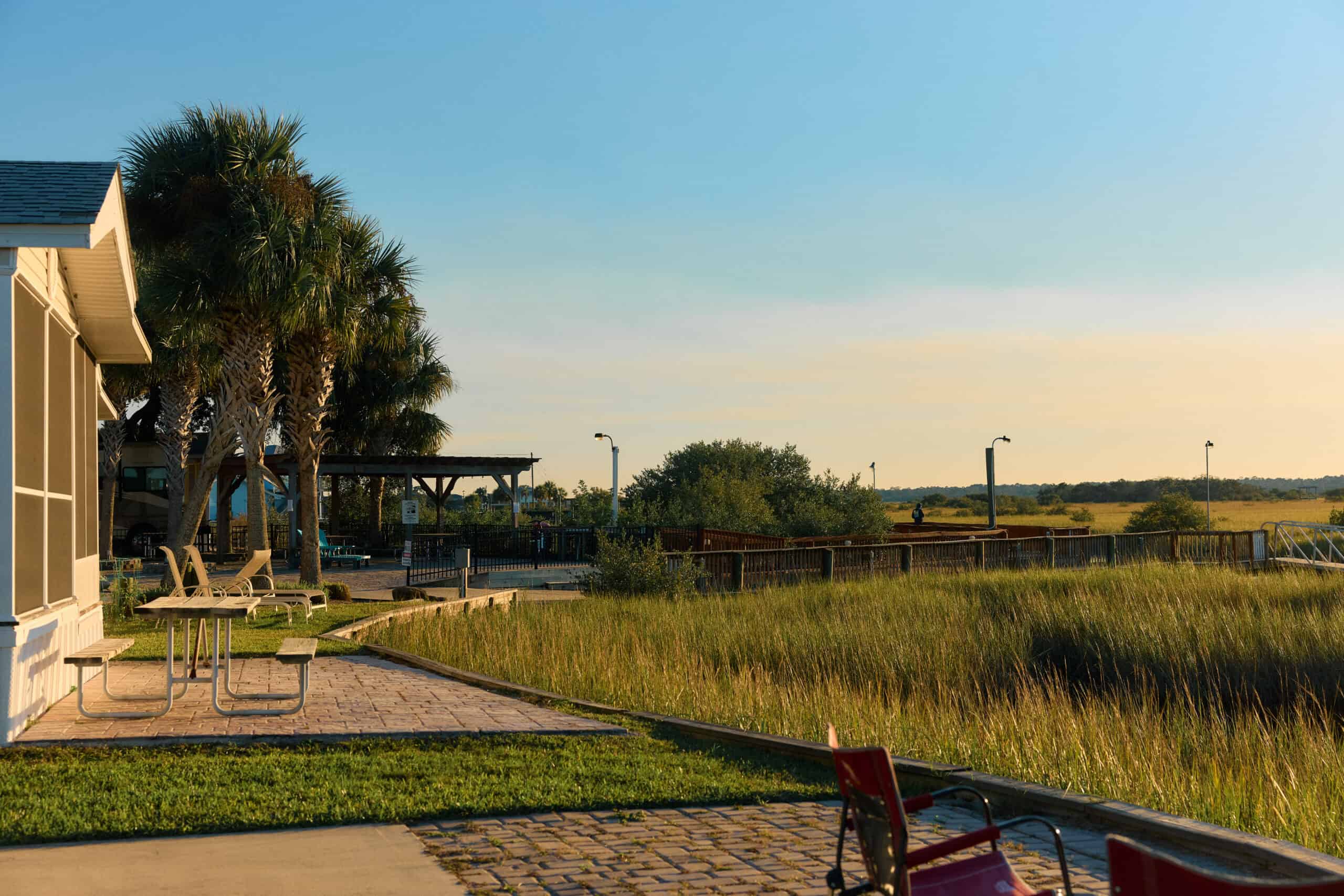 A patio with picnic tables overlooks a grassy marsh area, palm trees, and a pavilion under a clear blue sky in late afternoon sunlight.