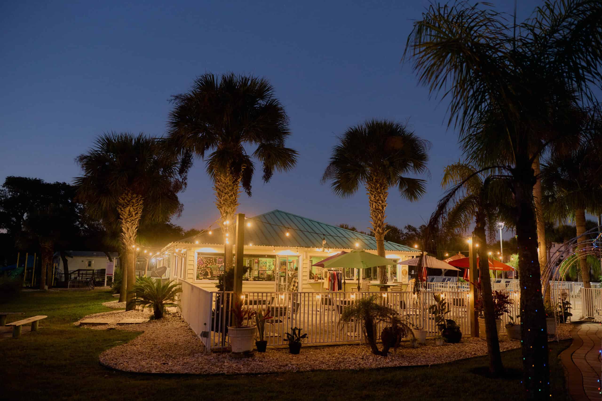 Outdoor restaurant with string lights and colorful umbrellas, surrounded by palm trees and a white fence, photographed at dusk.