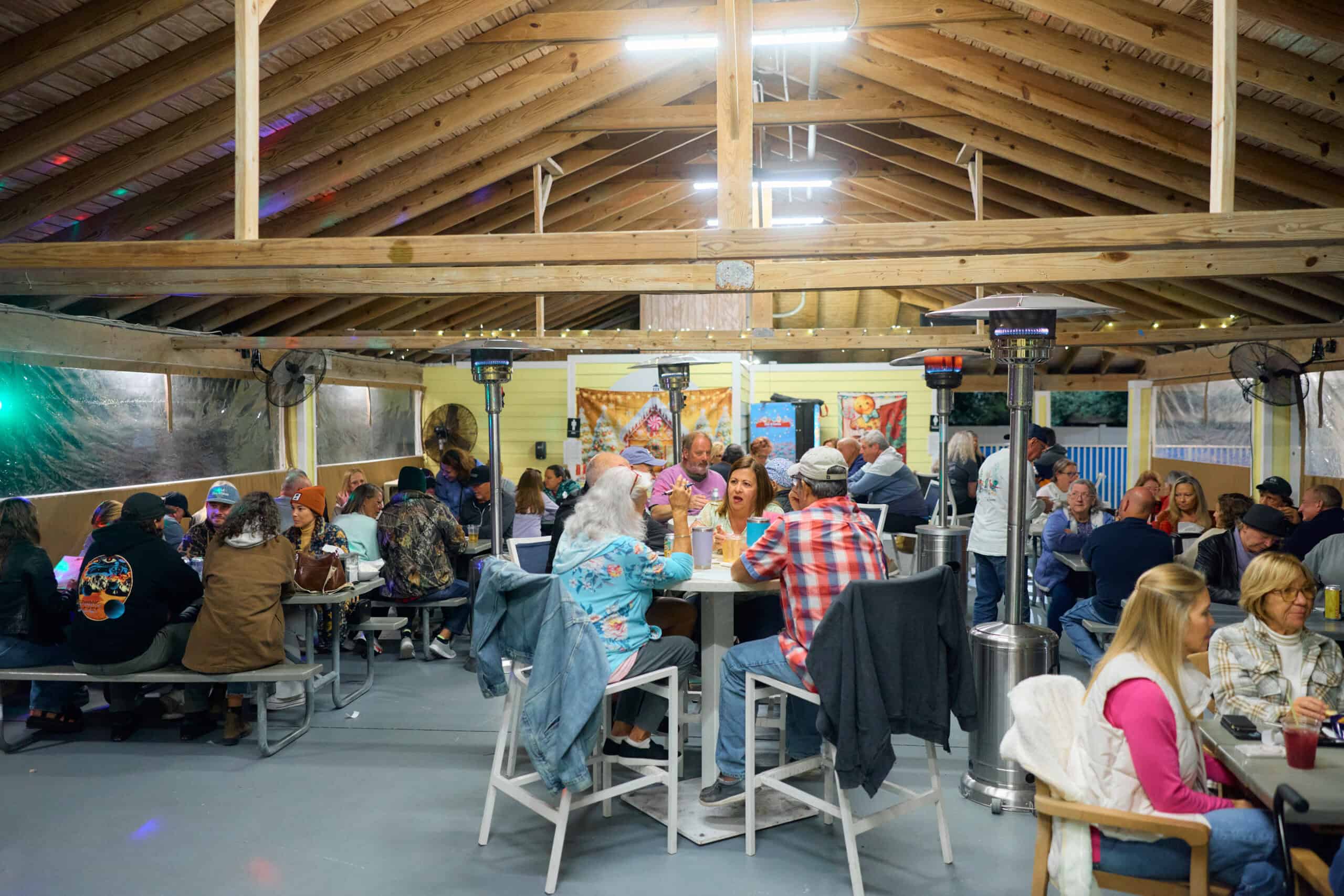 A large group of people sit at tables inside a covered outdoor dining area with a wooden ceiling, eating, drinking, and conversing.