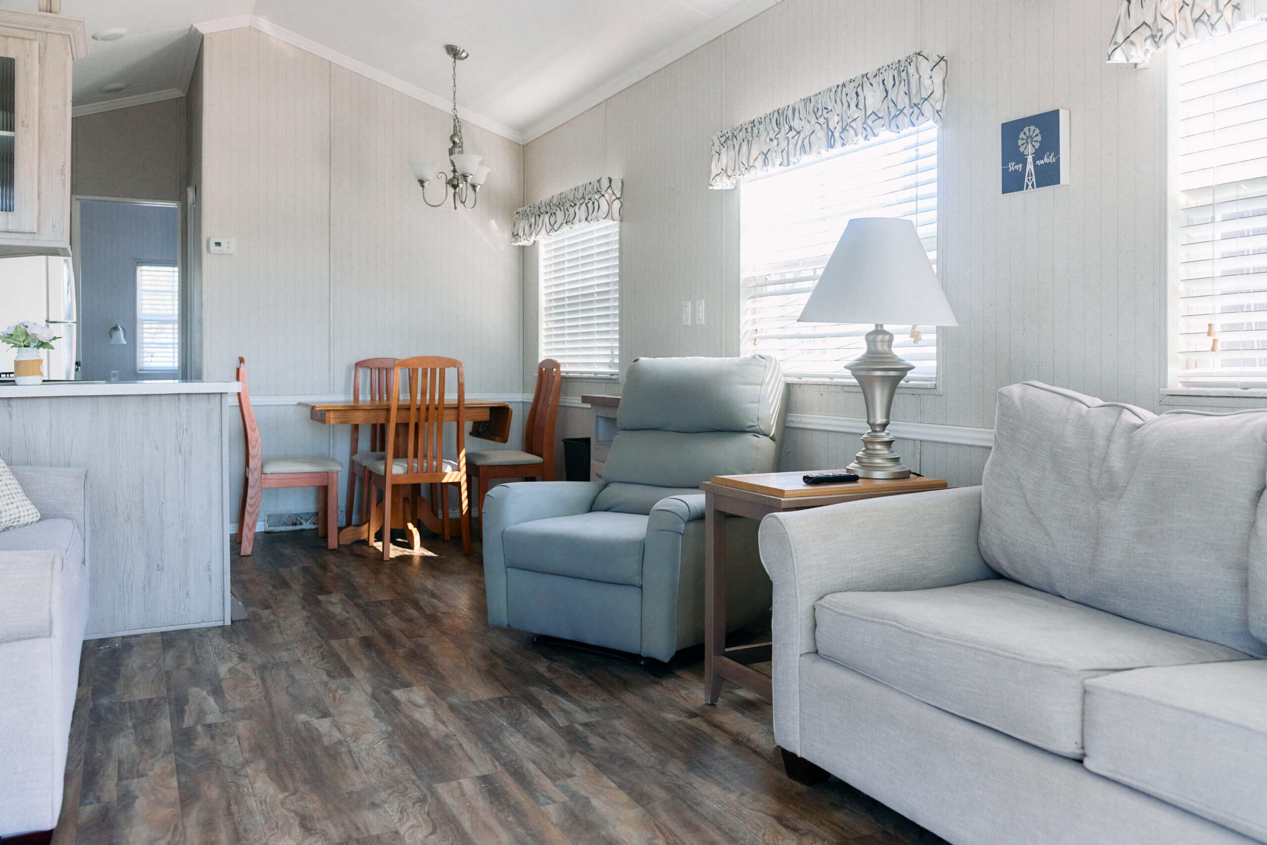 Living room with light gray sofa, recliner, side table with lamp, and dining area with wooden table and chairs; bright daylight streams through multiple windows.