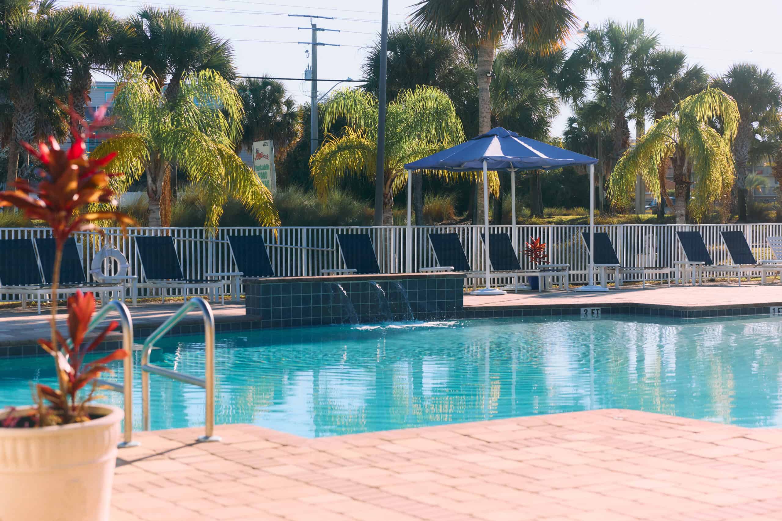 Outdoor swimming pool with blue lounge chairs and a blue umbrella, surrounded by palm trees and a white fence on a sunny day.