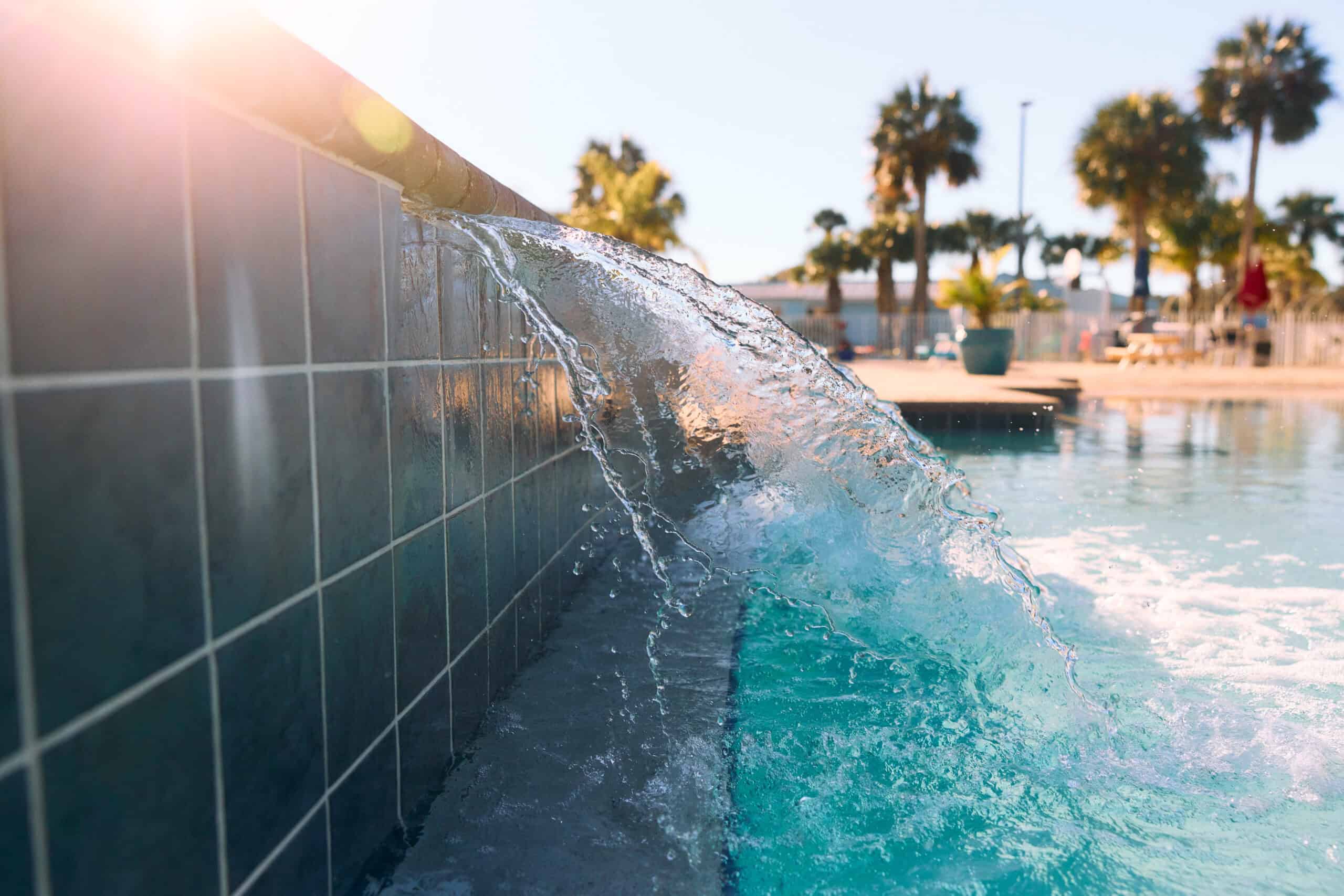Water flows over the edge of a tiled pool into clear blue water, with palm trees and bright sunlight in the background.