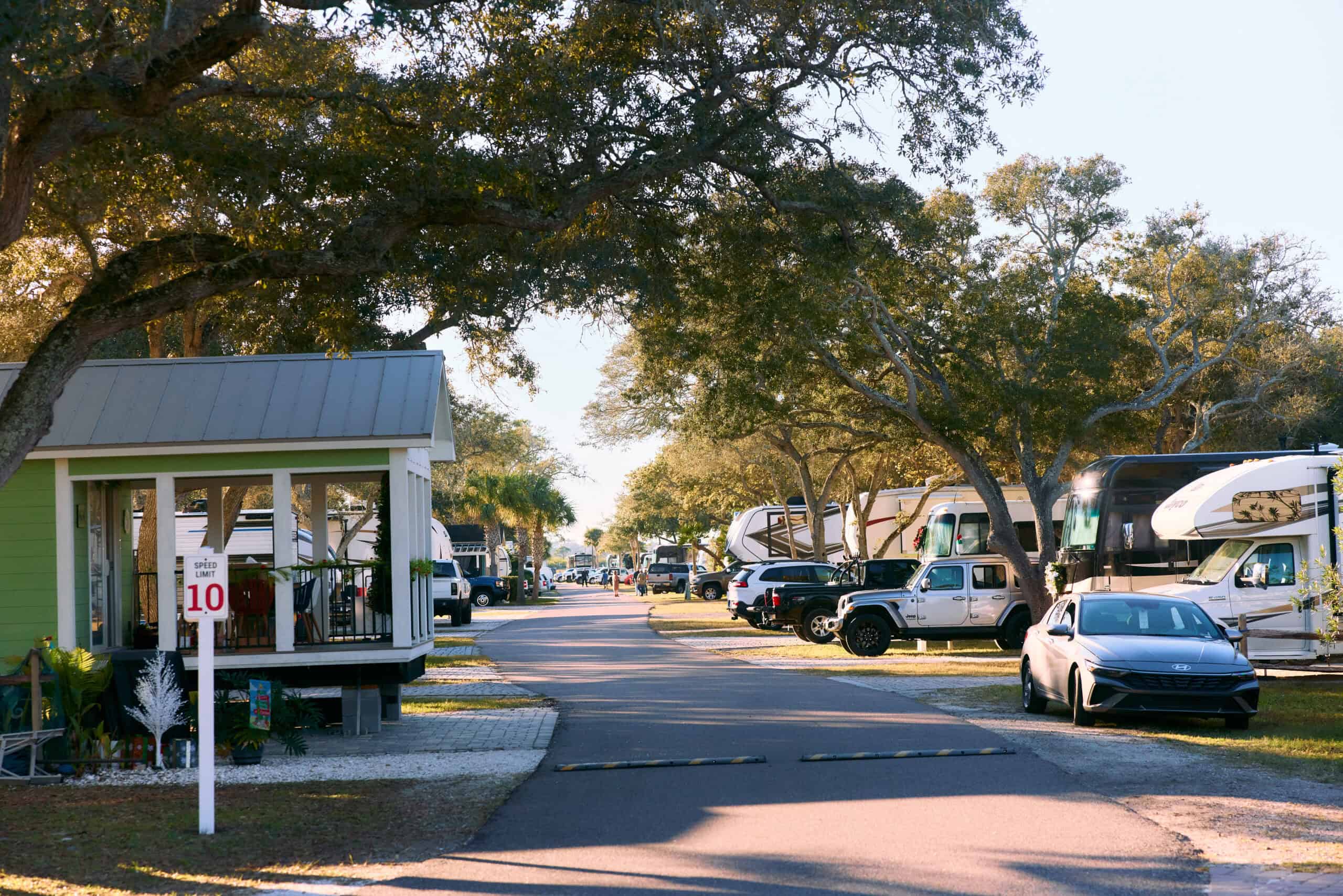 A paved road runs through an RV park with parked cars and motorhomes on both sides, shaded by large trees and a speed limit sign visible in the foreground.