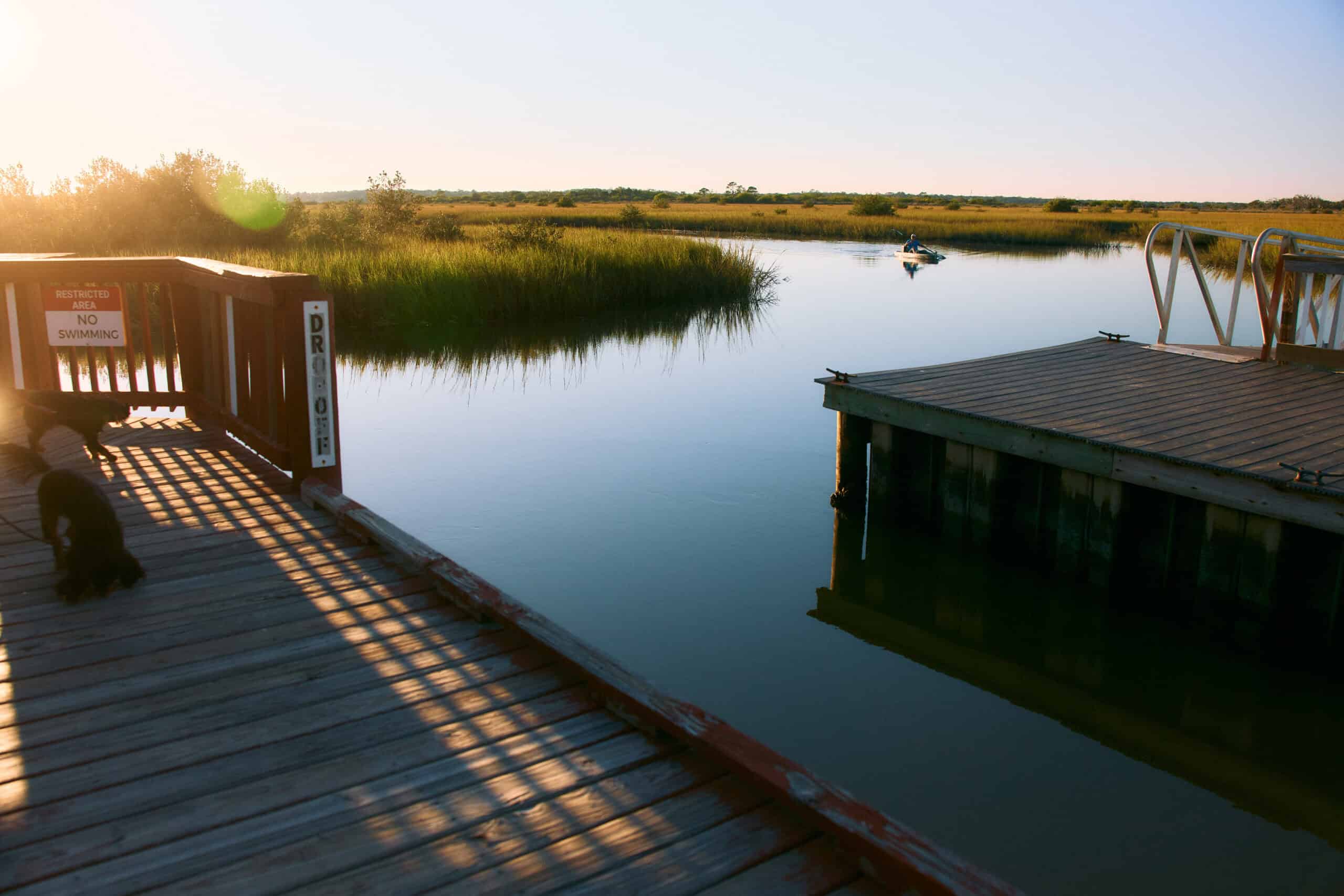 A calm river bordered by grassy banks, with a small dock on the right and a person kayaking in the distance. Sunlight casts shadows on the wooden deck in the foreground.