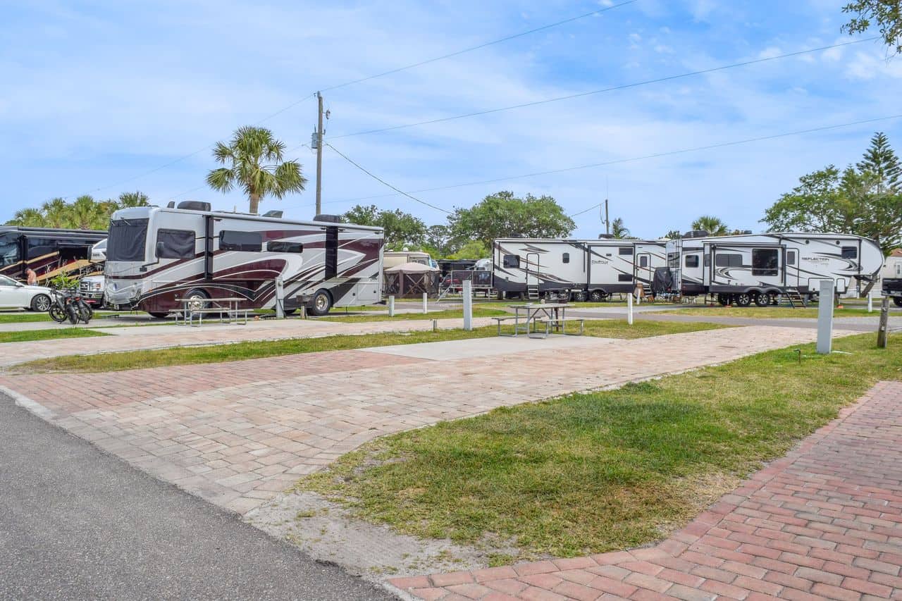 Several RVs and trailers are parked in marked spots at an RV campground, with picnic tables, grass areas, and palm trees visible under a partly cloudy sky.