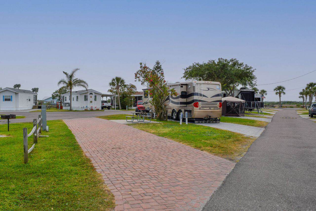 A large RV is parked on a brick driveway beside a picnic table in a campground with palm trees and nearby cabins under a clear sky.
