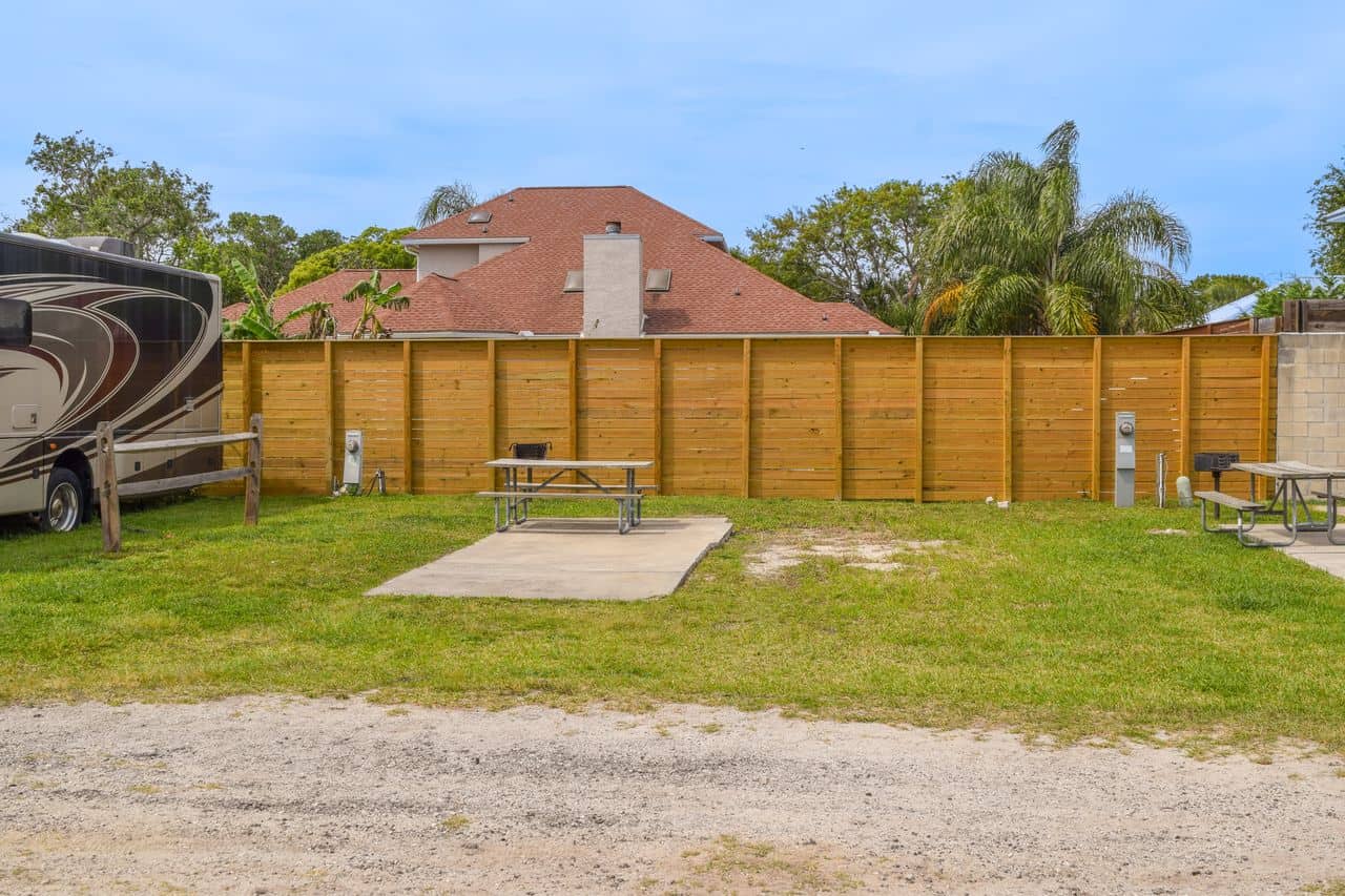 A grassy RV campsite with a concrete pad, picnic table, and wooden privacy fence; a house and palm trees are visible in the background.