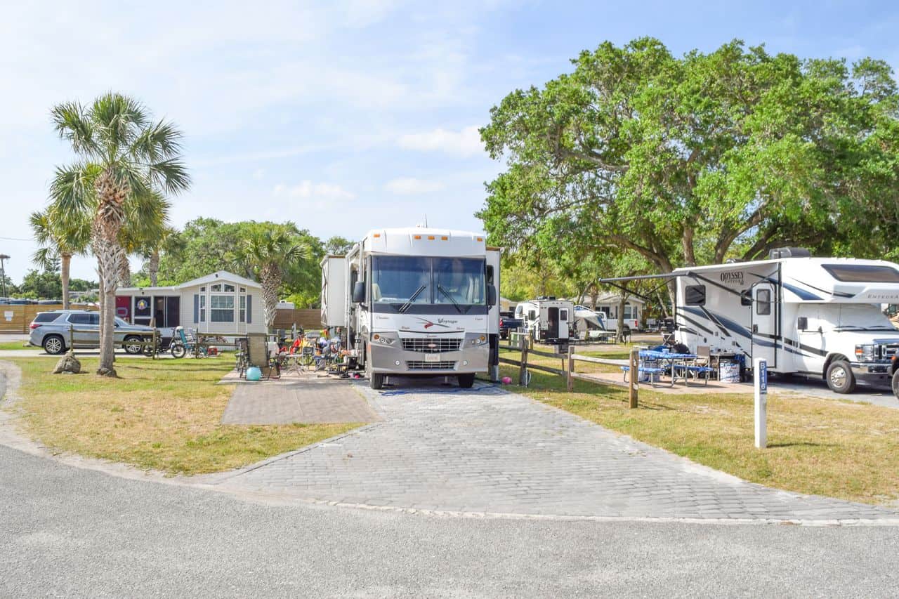 Three RVs are parked at a campground with trees, picnic tables, and outdoor seating visible under a sunny sky.