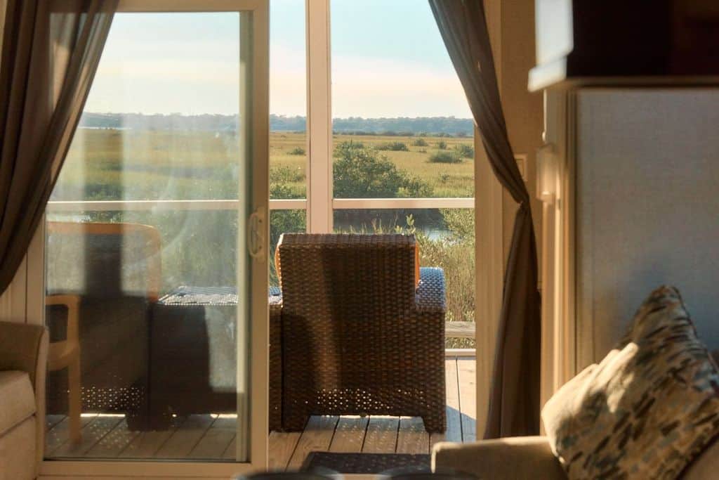 A wicker chair and table sit on a balcony outside a sliding glass door, overlooking a grassy landscape under a clear sky.