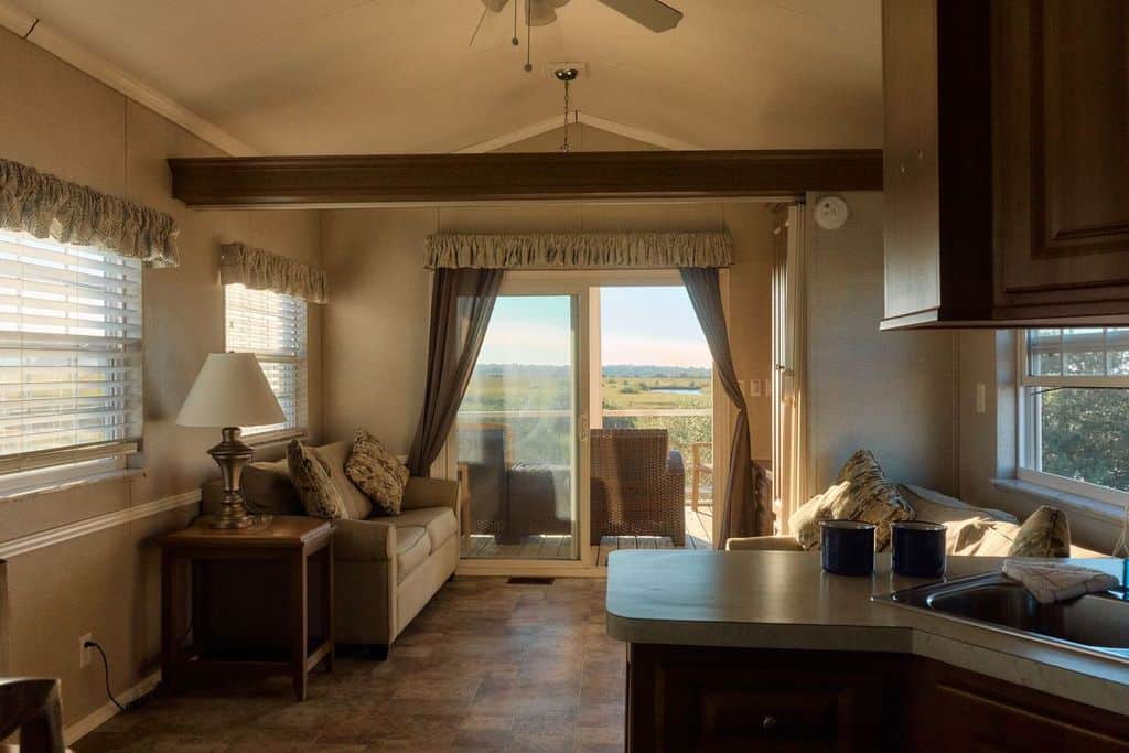 A cozy living room with two sofas, a side table with a lamp, and large glass doors opening to a patio with outdoor chairs and a view of open fields.