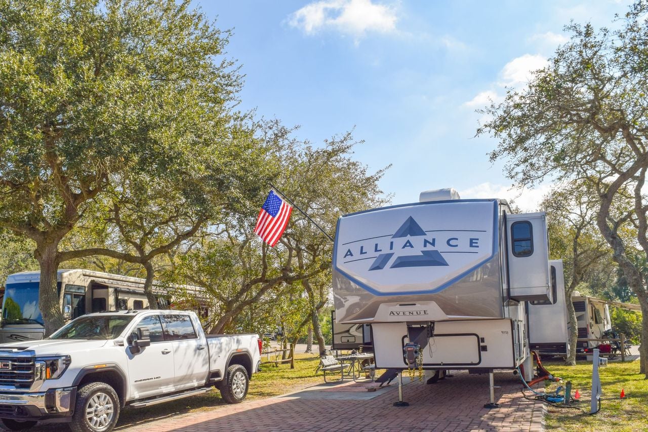 A large Alliance Avenue fifth-wheel RV is parked at a campsite beside a white pickup truck, with an American flag and trees in the background.