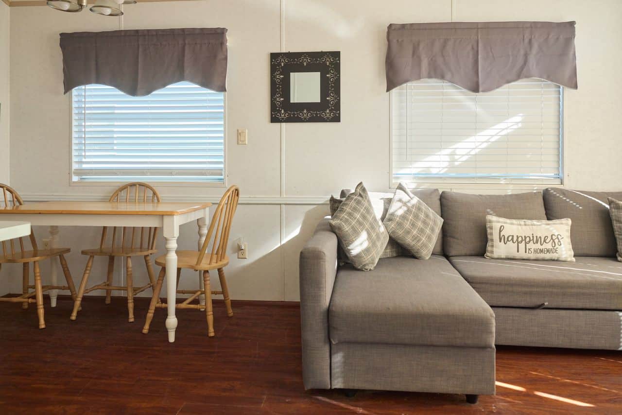 A small dining table with four wooden chairs sits beside a gray sectional sofa with plaid pillows and a "happiness is homemade" cushion in a bright room.
