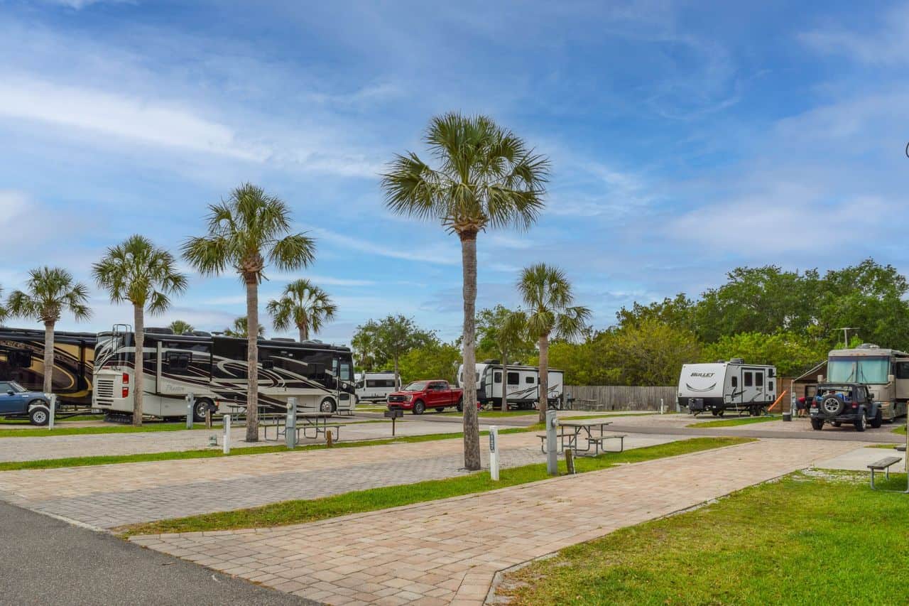 Several RVs and a red car are parked at a paved RV park with palm trees and grassy areas under a partly cloudy sky.