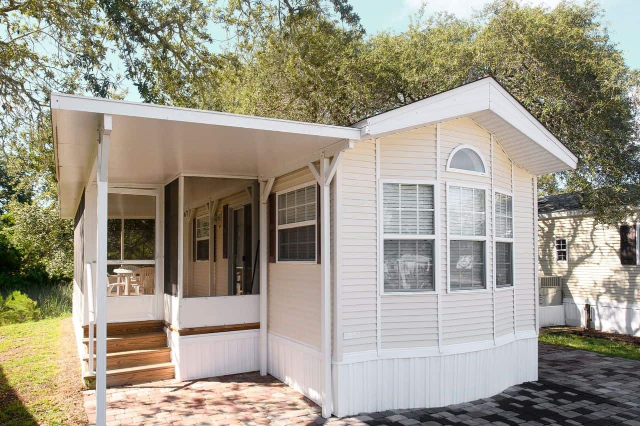 A small, white manufactured home with a covered porch, surrounded by trees and set on a brick-paved area.