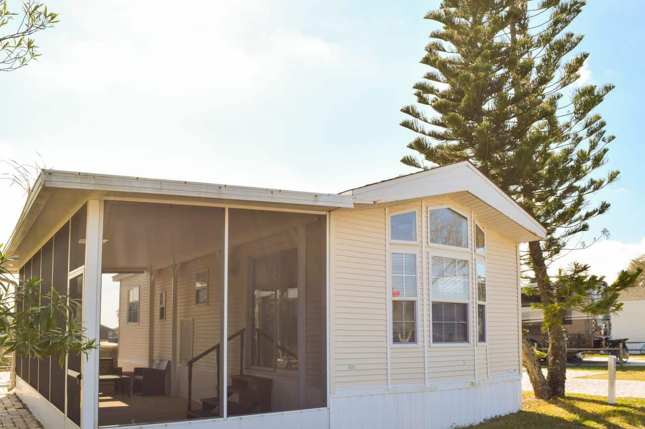 Single-story manufactured home with tan siding, large front windows, and a screened porch; a tall pine tree stands beside the house.