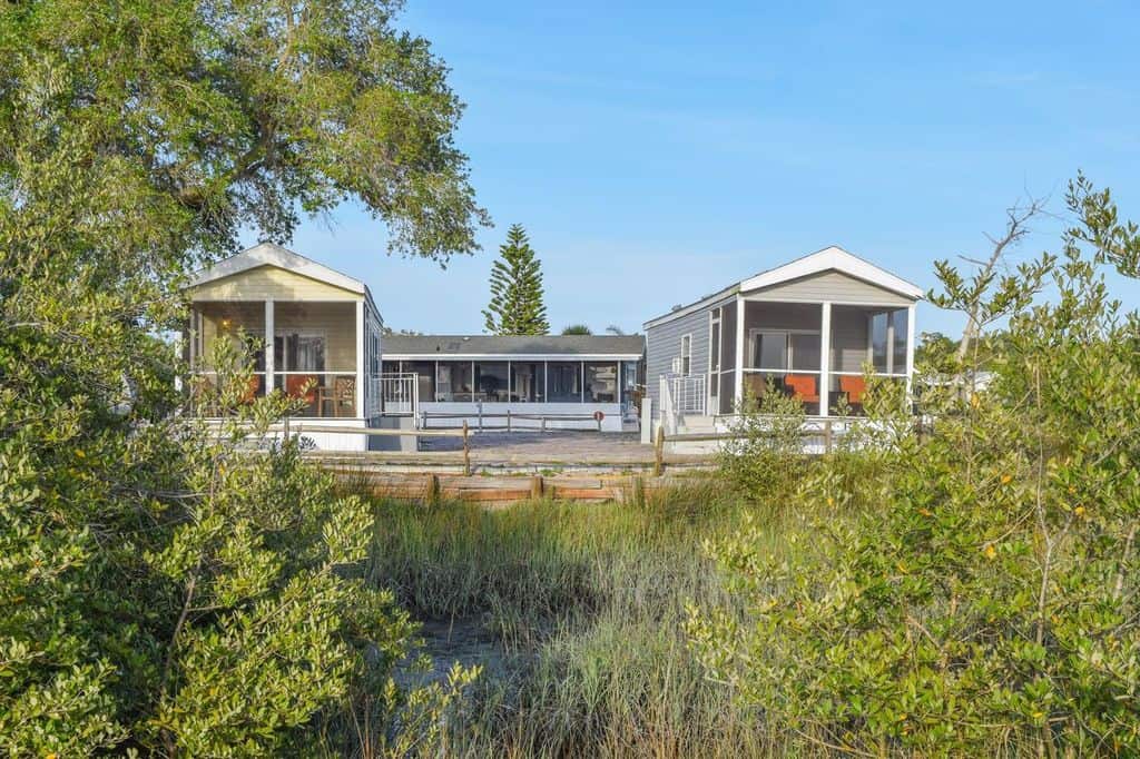 Two white single-story houses with screened porches and a central connecting building, surrounded by greenery and tall grass under a clear blue sky.