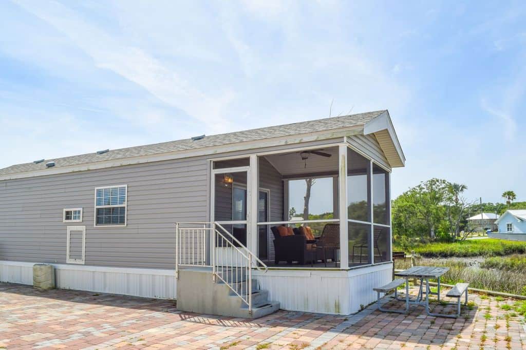 Gray manufactured home with a screened porch, outdoor steps, patio area, and picnic table set beside a grassy area under a partly cloudy sky.