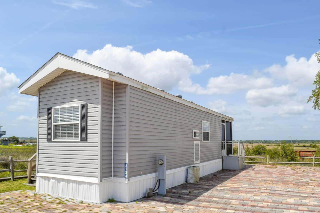 A gray manufactured home with white trim sits on a paved lot under a blue sky with scattered clouds, surrounded by grass and a wooden fence.