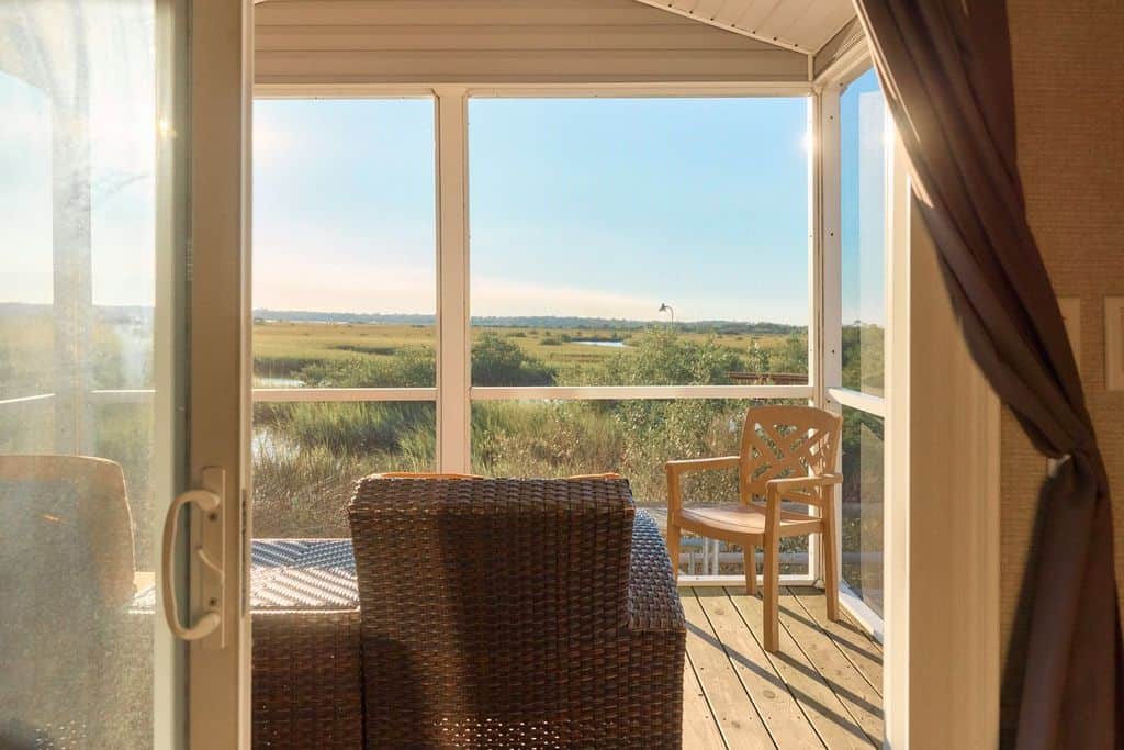 View from inside a room looking out onto a sunlit porch with wicker furniture, overlooking grassy wetlands and a distant body of water.