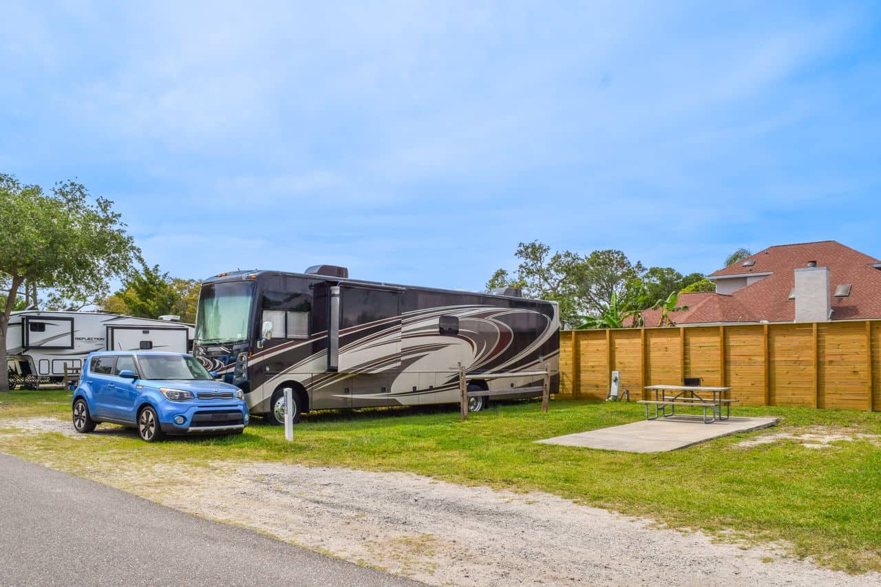 A large motorhome and a blue car parked at an RV campsite with a concrete pad, picnic table, and wooden privacy fence on a clear day.