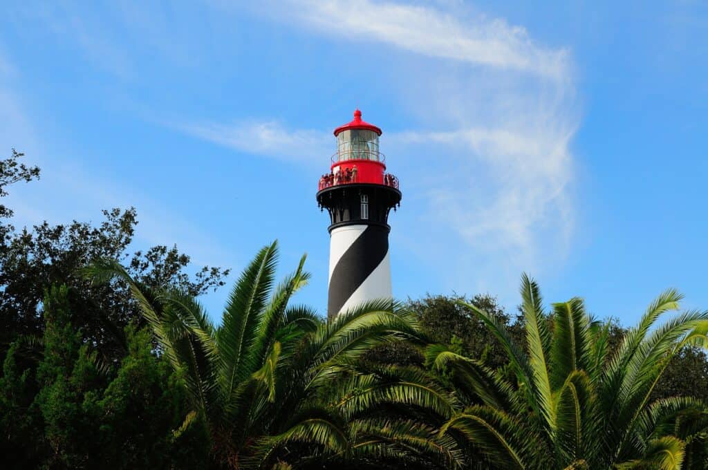 A black and white striped lighthouse with a red top rises above green palm trees under a blue sky with wispy clouds.