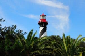 A black and white striped lighthouse with a red top rises above green palm trees under a blue sky with wispy clouds.