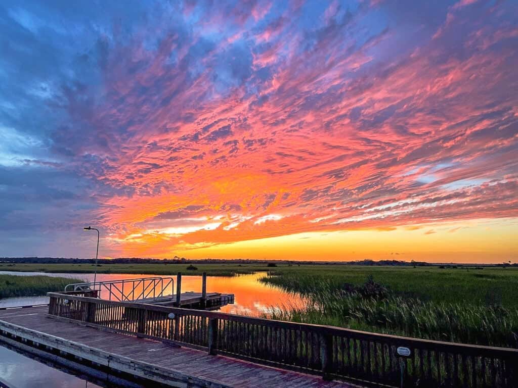 Wooden dock by a marsh with tall grass at sunset, featuring vibrant orange, pink, and blue clouds filling the sky.