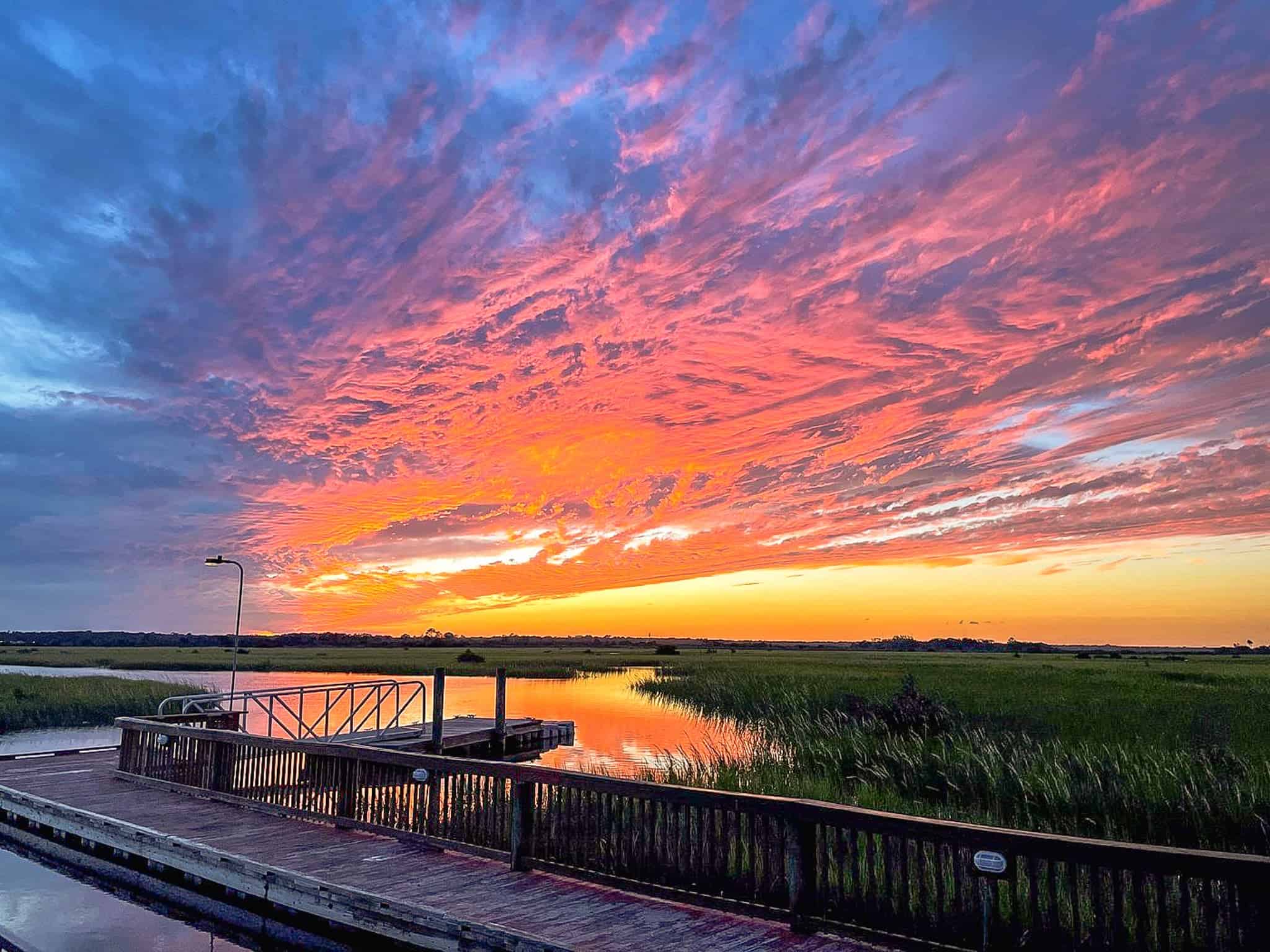 Wooden dock by a marsh with tall grass at sunset, featuring vibrant orange, pink, and blue clouds filling the sky.