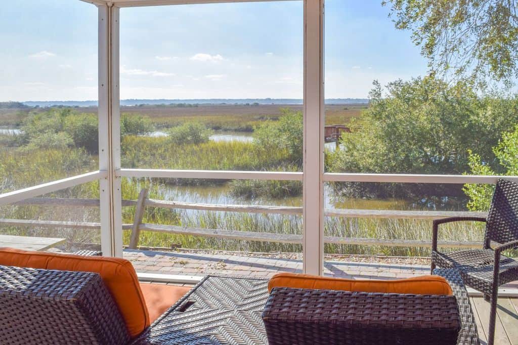 Screened porch with wicker chairs and orange cushions overlooks a marsh with tall grass, water, and distant trees under a clear blue sky.