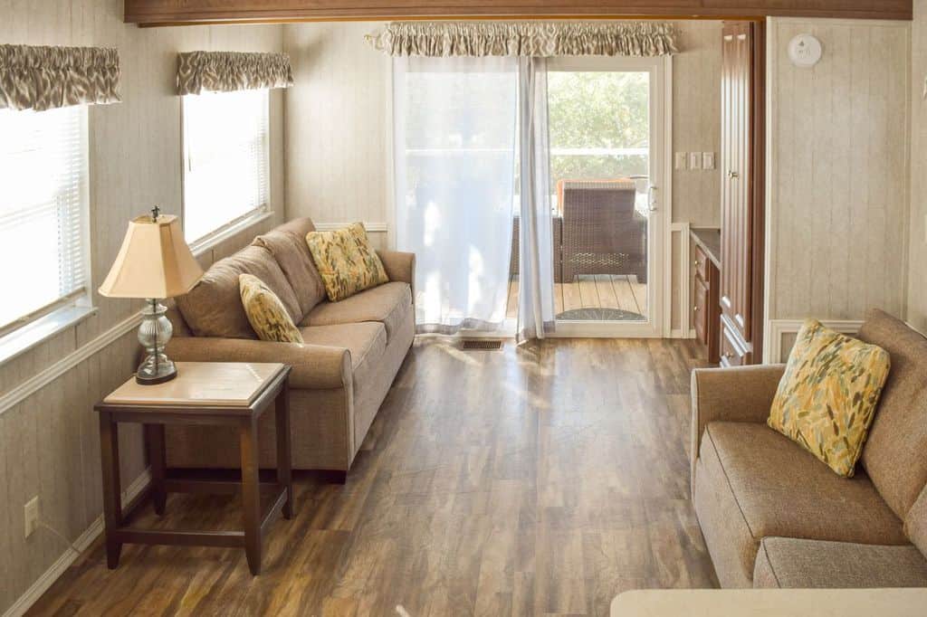 A bright living room with two brown sofas, a side table with a lamp, large windows, wood flooring, and a sliding glass door leading to a sunlit deck with outdoor seating.