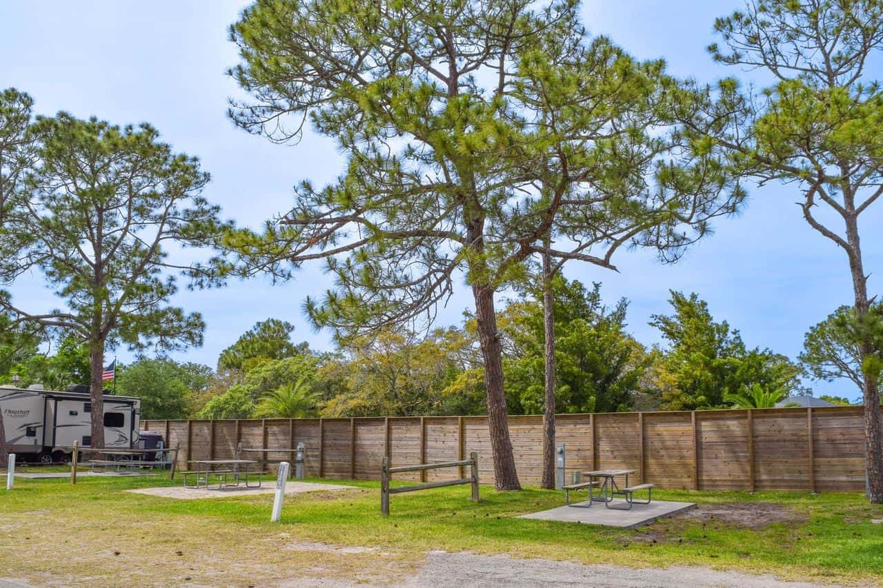 Two empty picnic tables and benches sit by a wooden fence, with large pine trees providing shade in an open campground area. An RV is partially visible on the left.