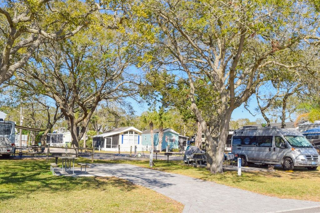RVs and small houses are arranged under large leafy trees in a sunny, grassy area with paved walkways and picnic tables.