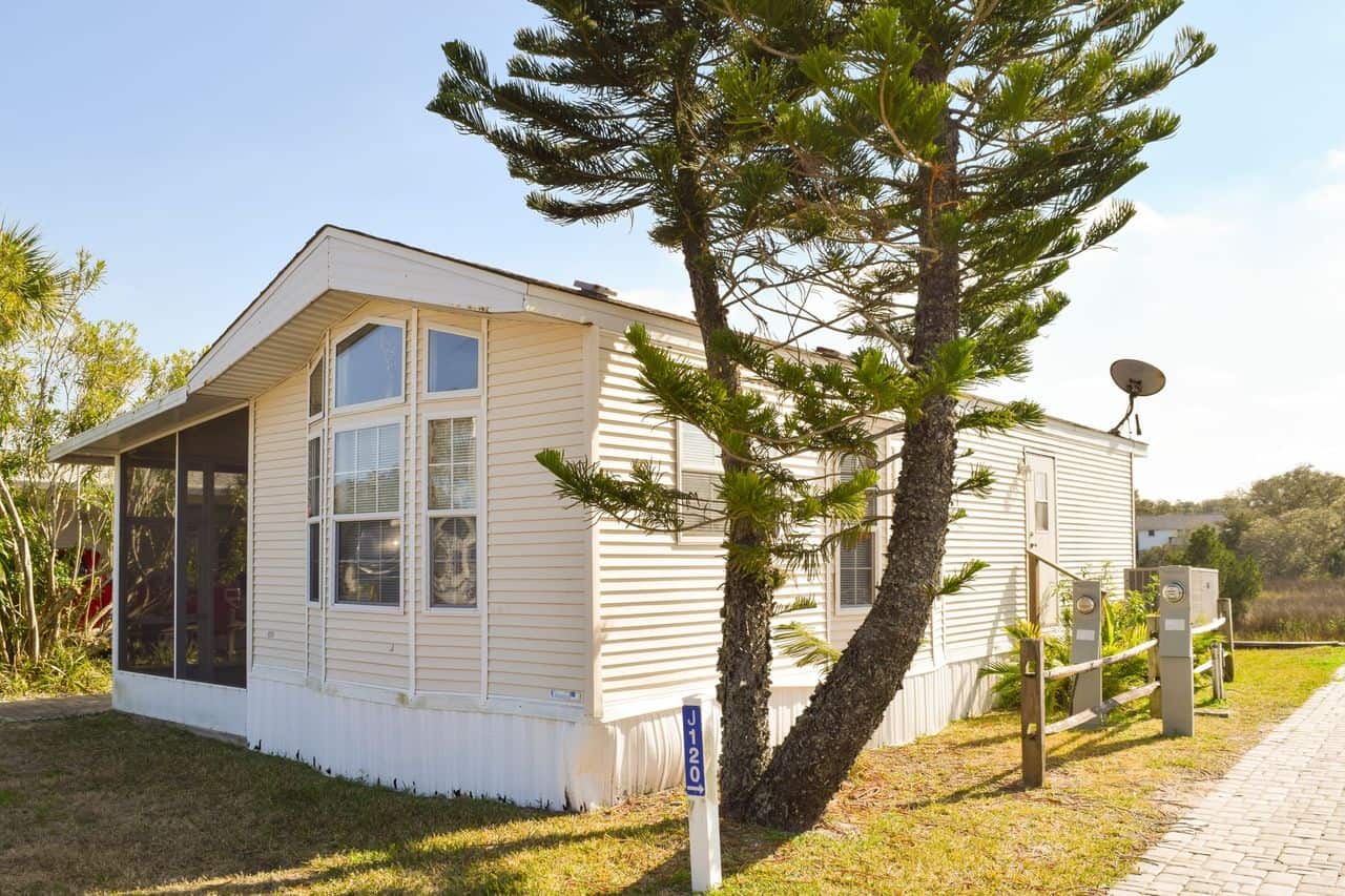 A beige manufactured home with large front windows, a screened porch, and a pine tree in the yard beside a paved walkway on a sunny day.