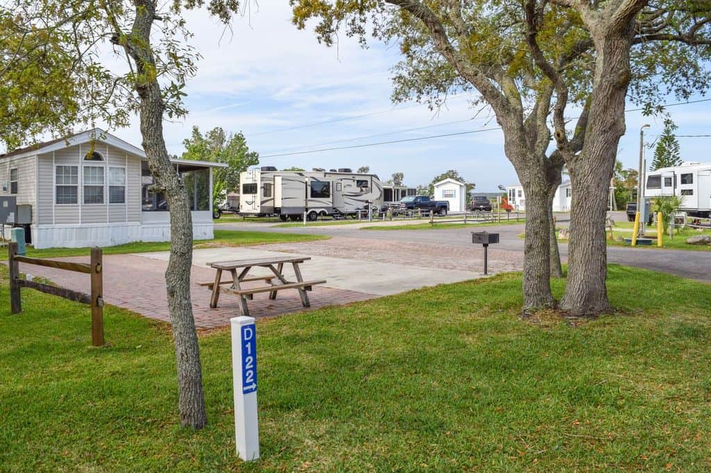 A paved RV site with a picnic table, grill, and trees, surrounded by grass. Several RVs and a small building are visible in the background. A blue marker labeled D122 is in the foreground.