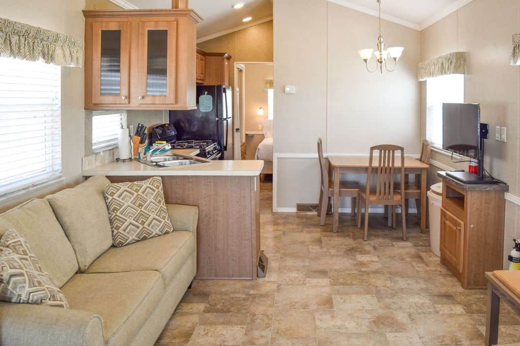 A compact living space with a beige sofa, small kitchen area, dining table with two chairs, TV on a stand, and neutral-colored decor.